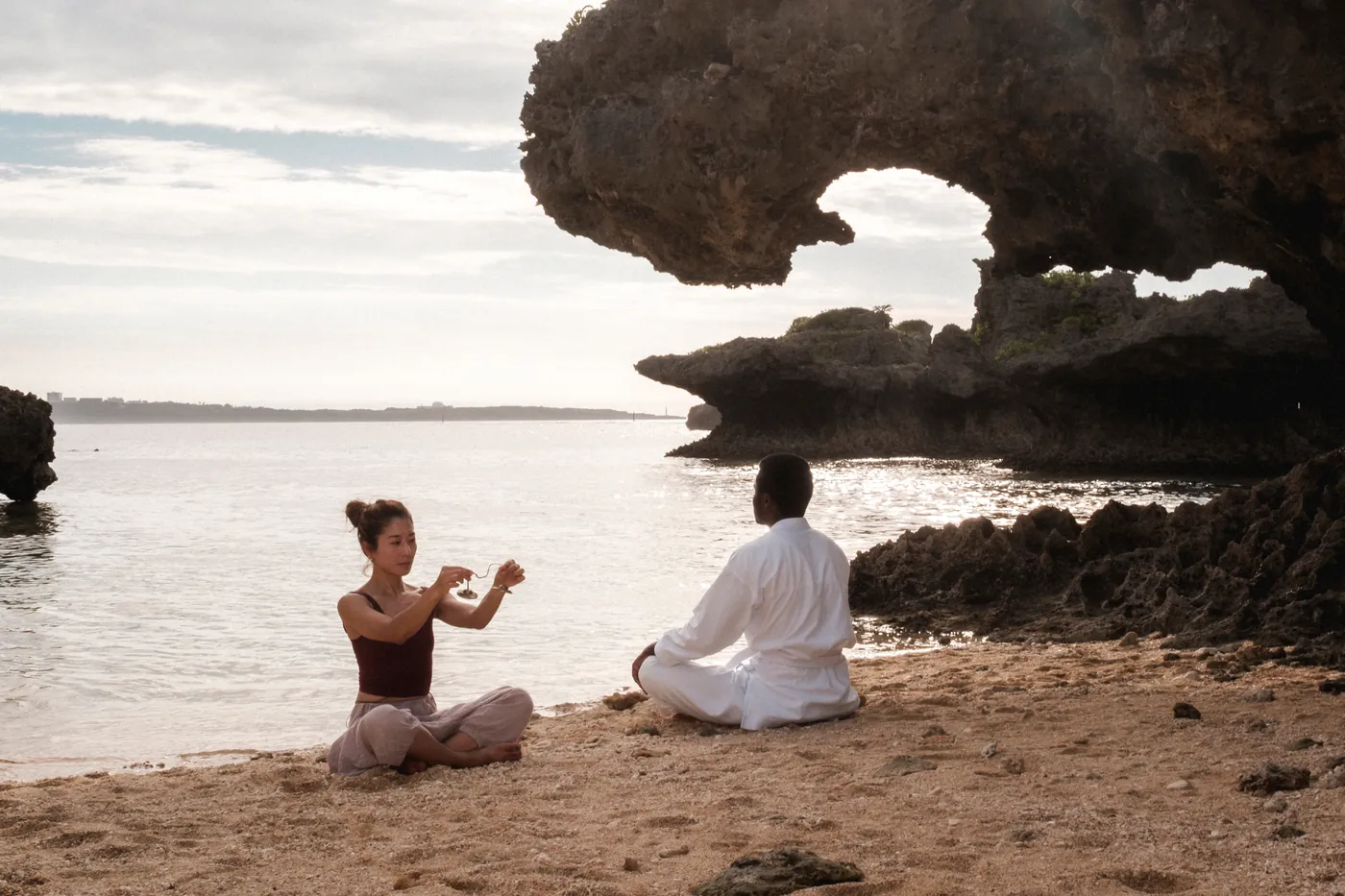 A woman and a karateka seated in quiet ceremony on a beach beneath a rock arch